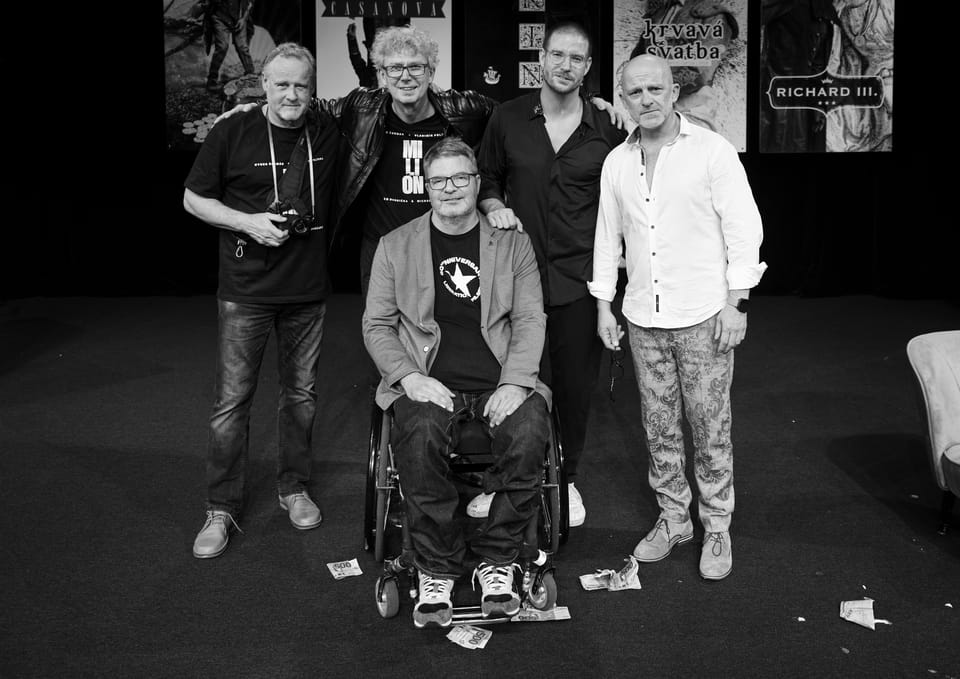 The company posing on stage before a wall of theatre posters, one of them in a wheelchair