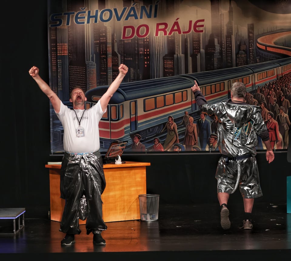 Two actors in silver costumes with arms raised before a painted backdrop