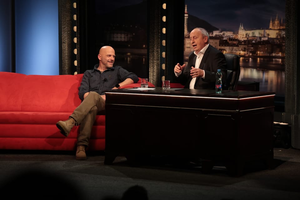 Two men on the red sofa of a television studio, a city panorama behind them