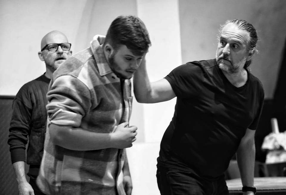 Three actors in motion on a bare stage, the front one leaning forward