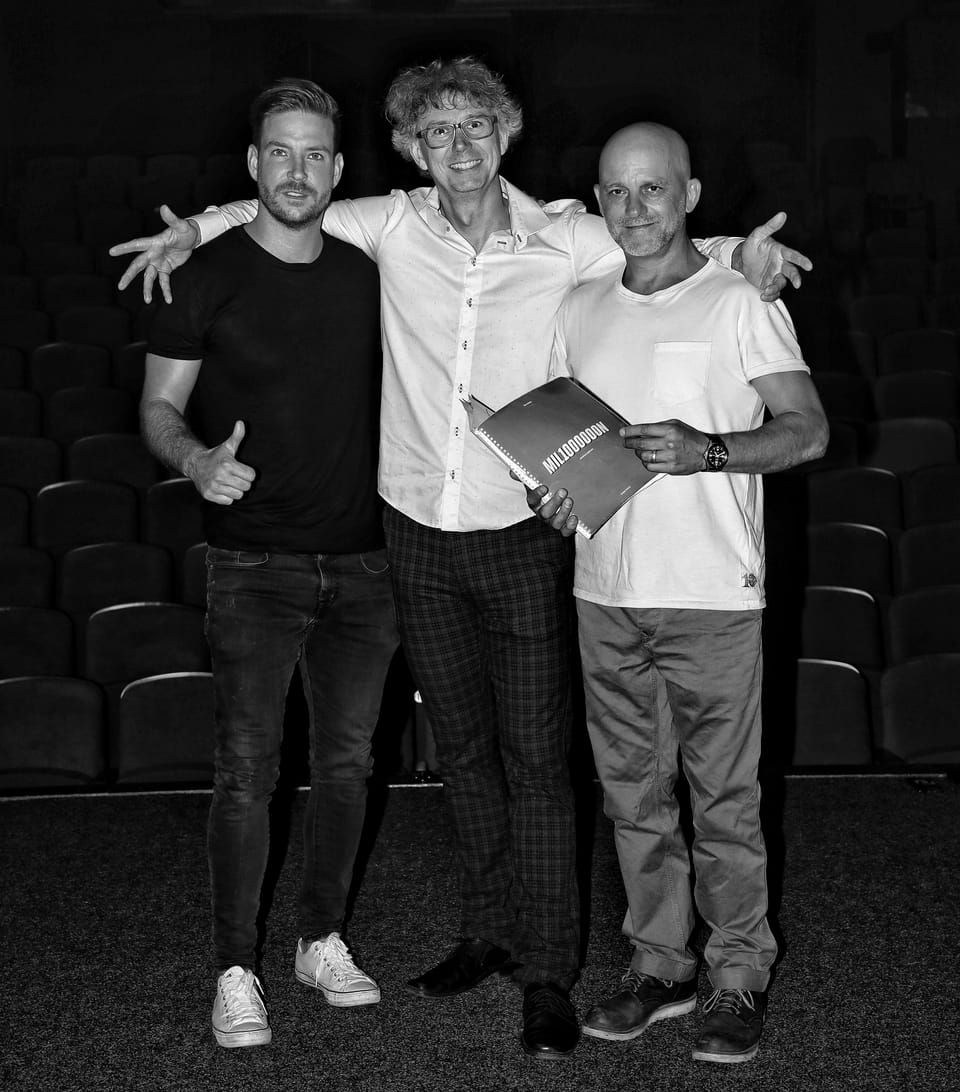 Three men in an empty auditorium, the middle one holding a book with arms wide