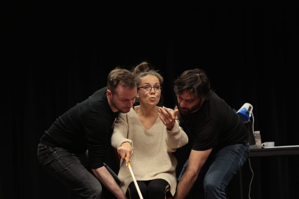 Three on stage, the woman at the centre holding a cane, both men leaning toward her