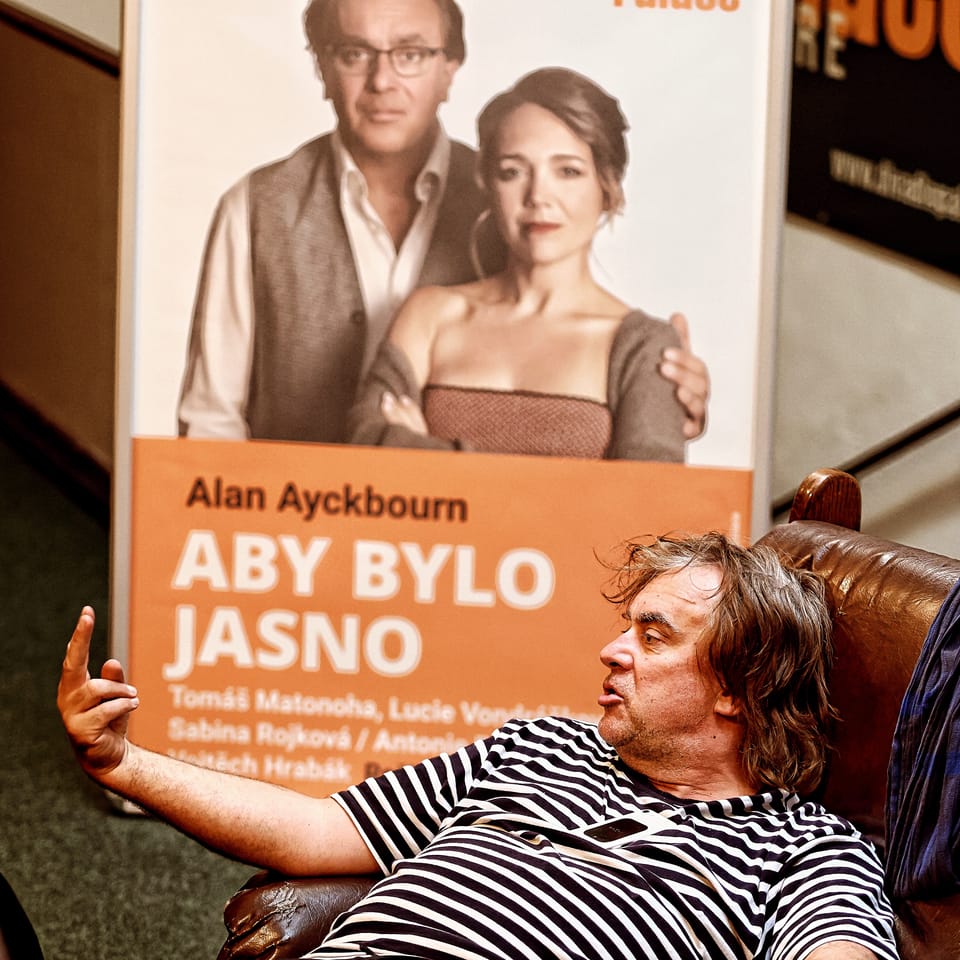 An actor sprawled in an armchair beside the poster of the play he is in