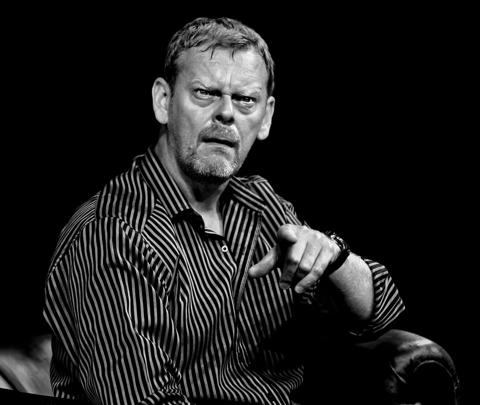 An actor in a striped shirt pointing into the house, brows drawn down