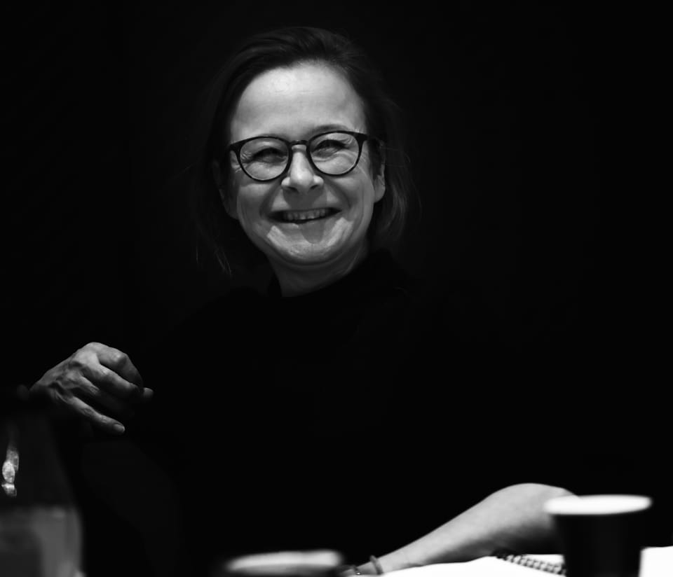 A woman in glasses smiling into the dark, the light finding only her face and hand