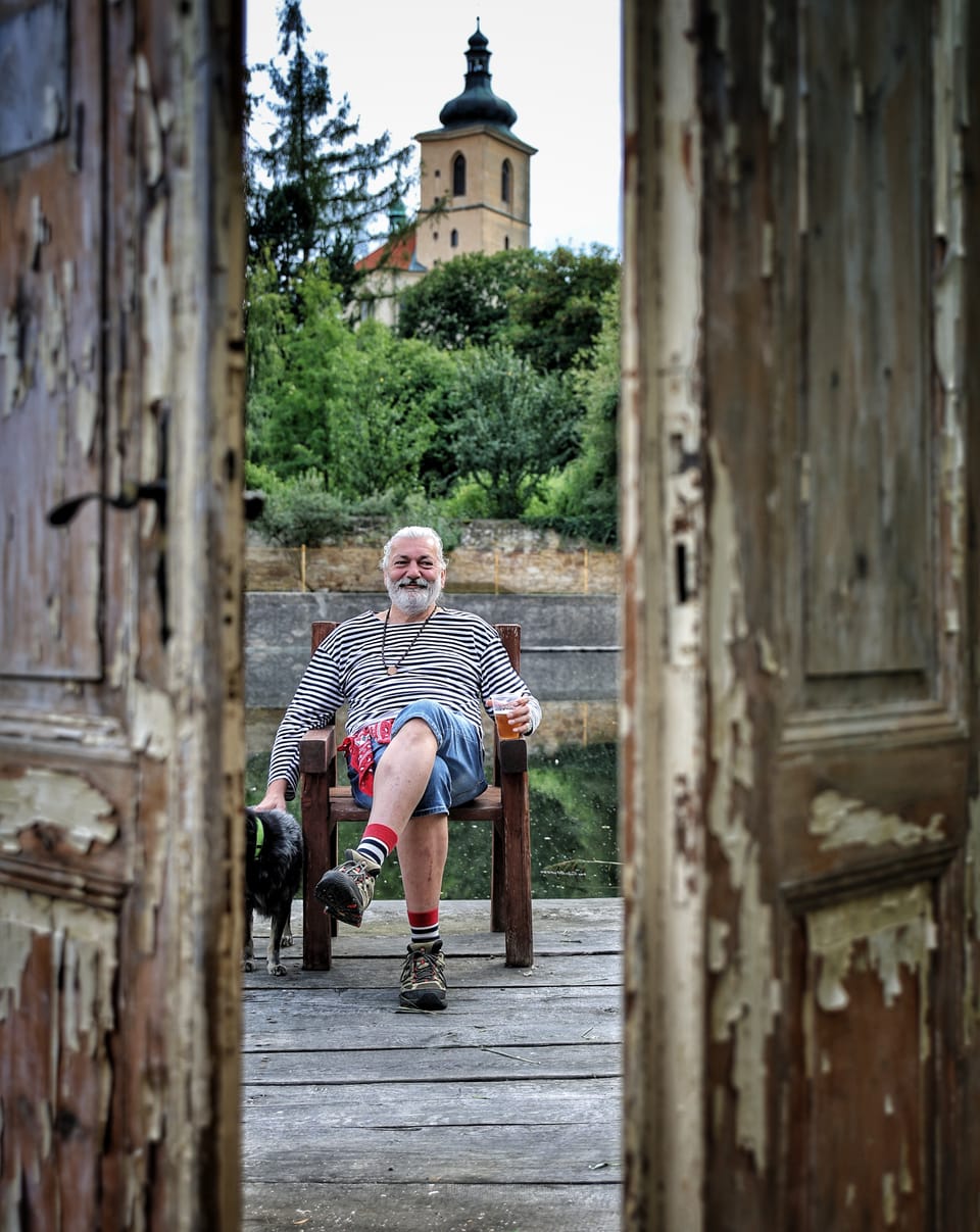 A man seated on a chair in a crumbling gateway, a church tower behind him