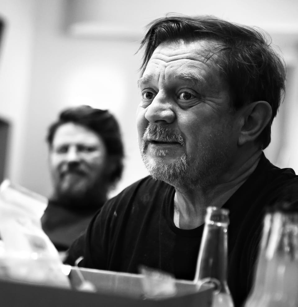 An older man with tousled hair at a table, another laughing behind him