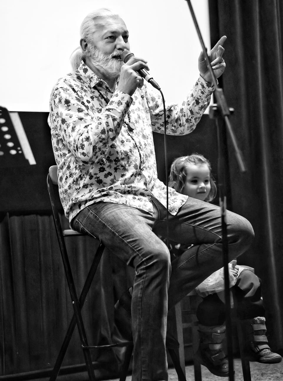 A bearded man with a microphone on a bar stool, a child sitting at his feet