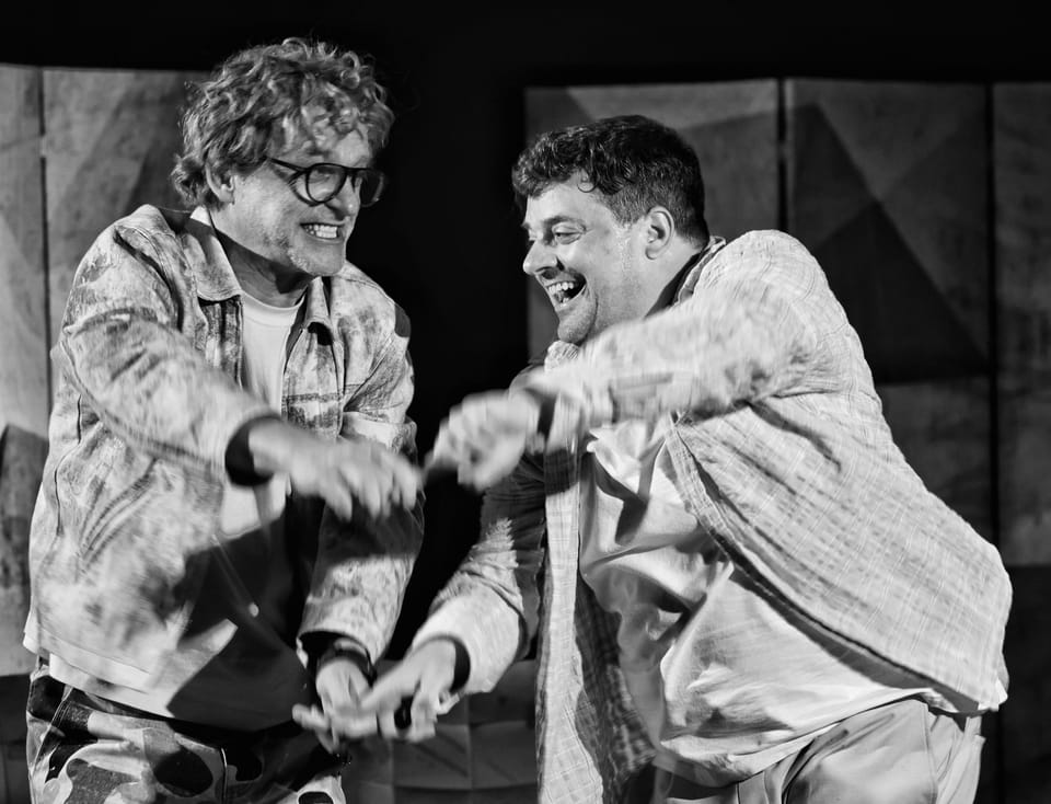 Two actors in dust-caked costumes, hands reaching toward each other