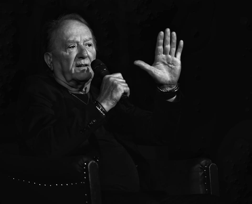 An older man at the microphone raising a palm as if stopping a question