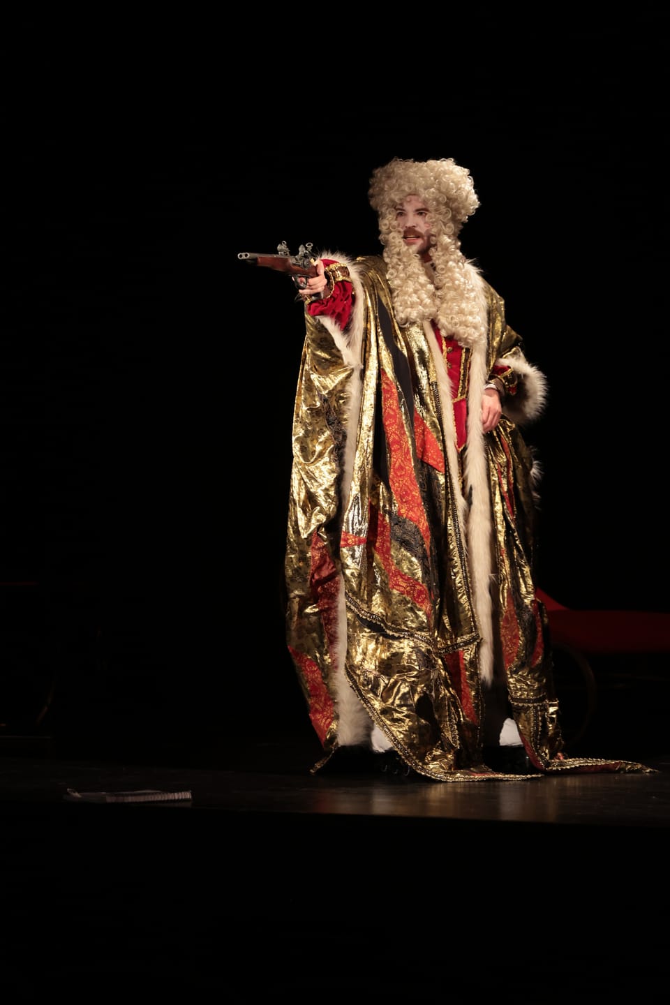 An actor in a gilded cloak and wig alone on the dark stage