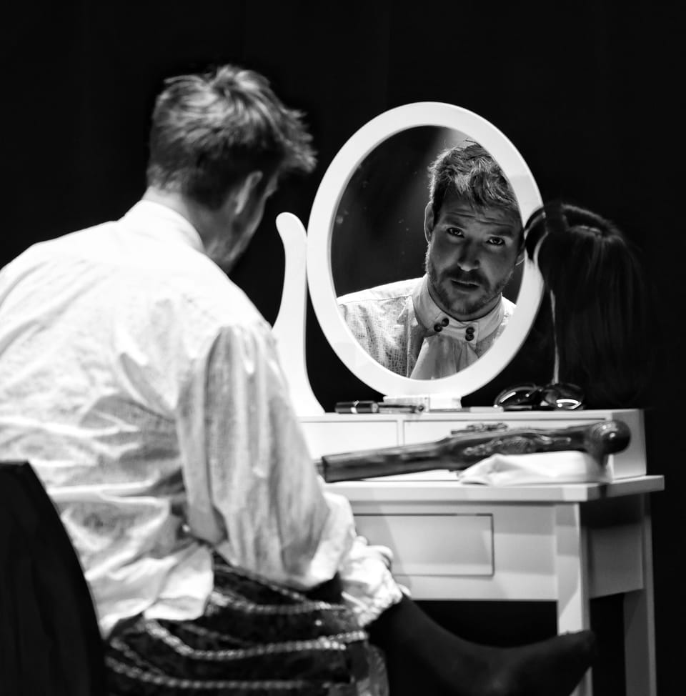An actor in costume at a dressing room mirror, his reflection looking back