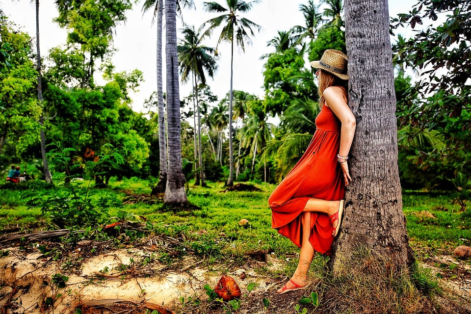 A woman in a red dress and straw hat leaning against a palm trunk