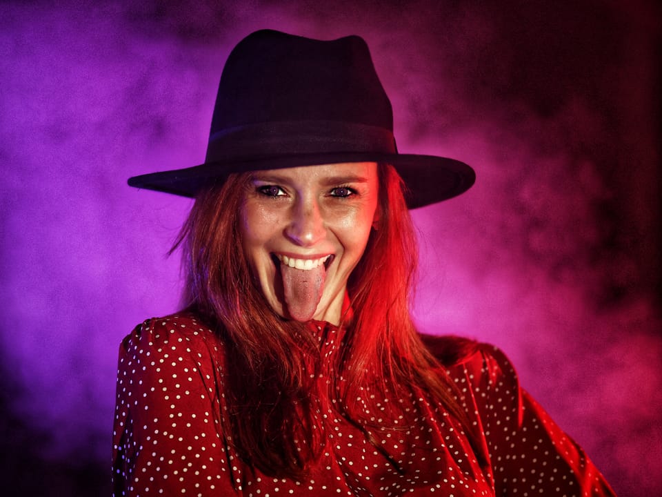 A glamour portrait of a red-haired woman in a black hat against magenta smoke