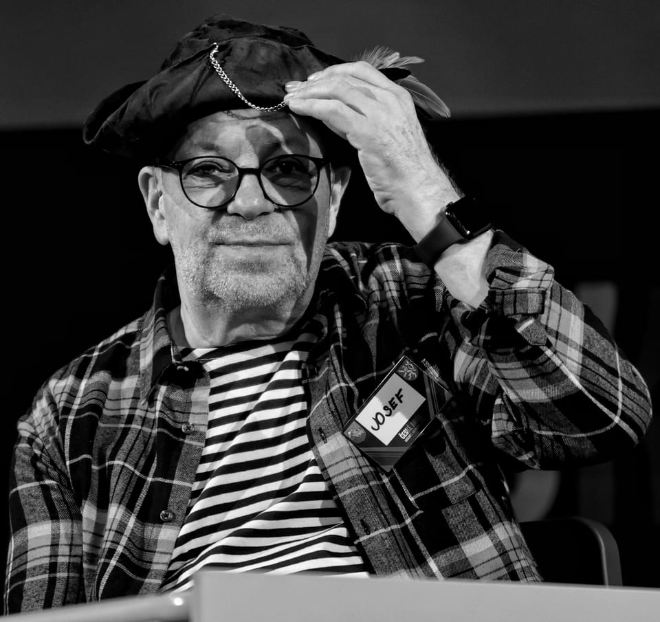 A man in a beret and glasses adjusting his hat, a name badge on his chest