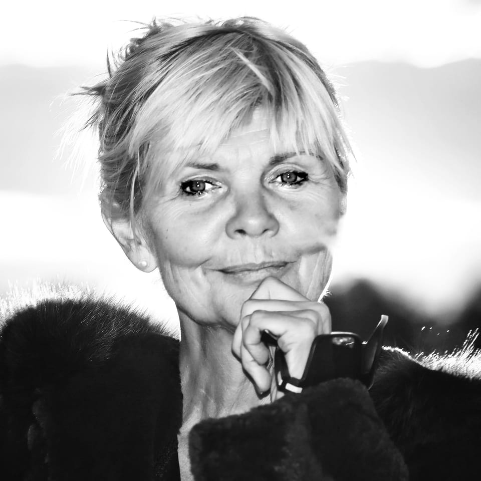 An older woman with short hair holding her glasses at her chin, bright landscape behind