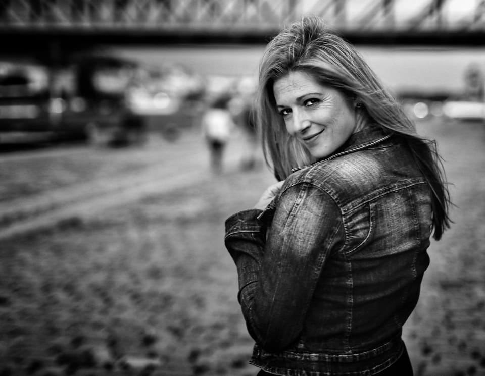 A woman in a denim jacket looking back, the steel of a bridge behind her