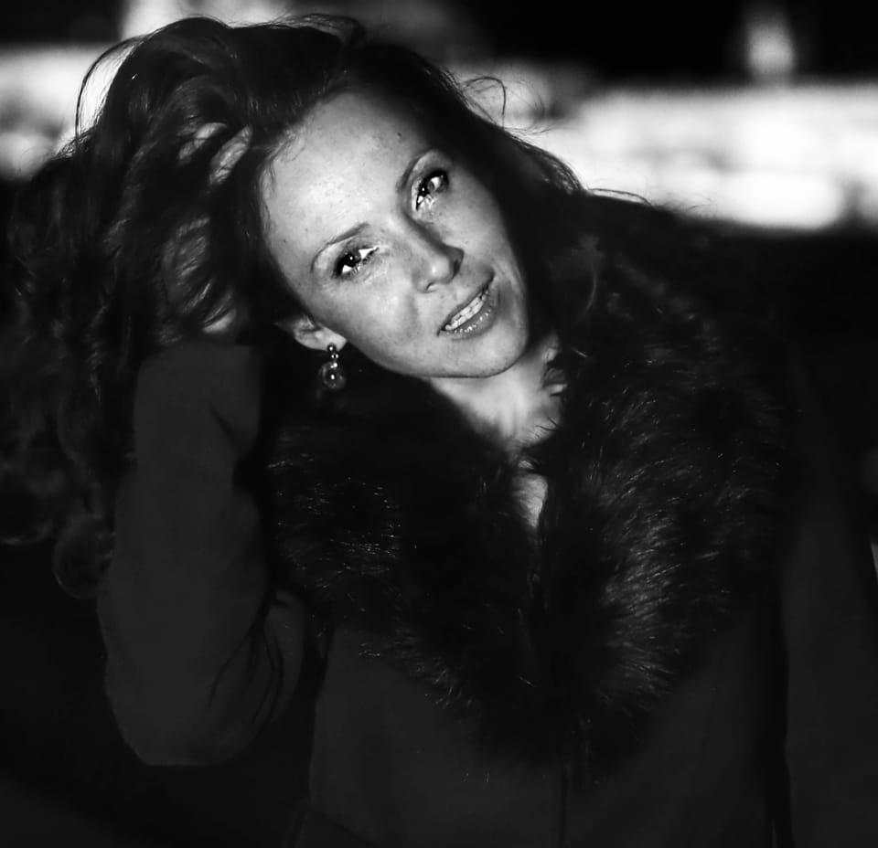 A woman with hands in her hair above a fur collar, a smile at the corner of her mouth