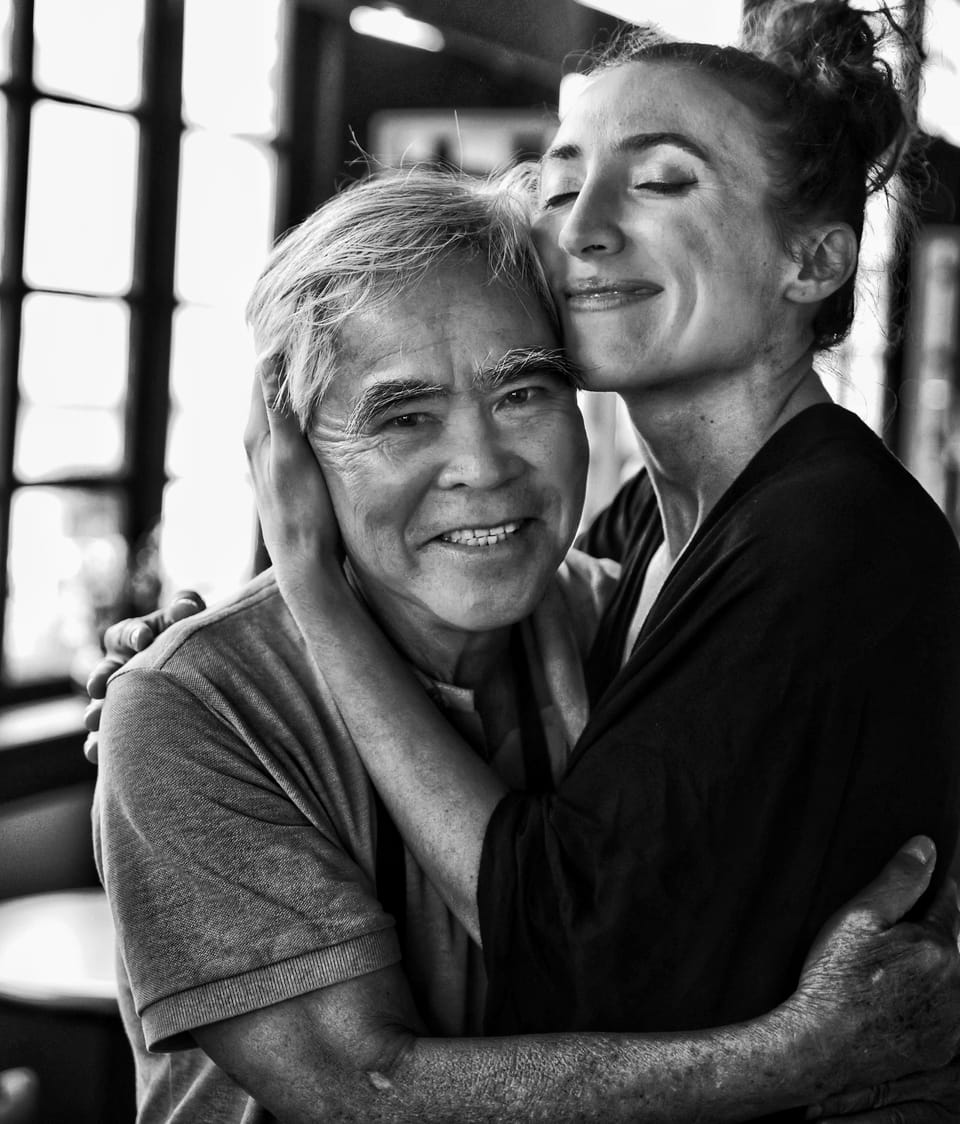 A black and white portrait of an older man and a woman embracing by a window