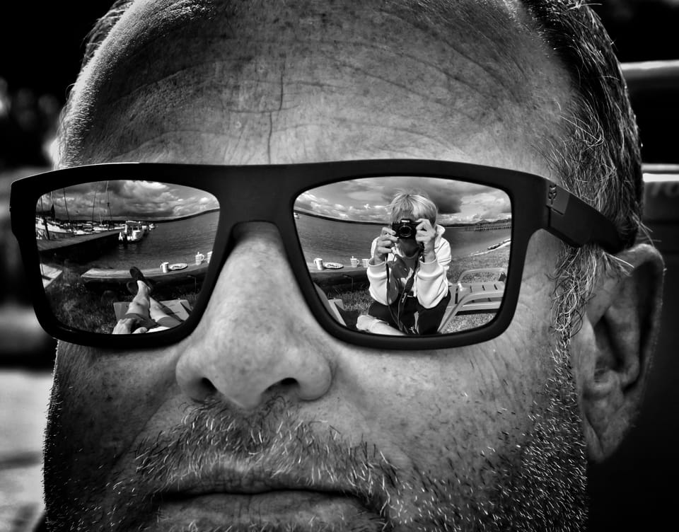 A detail of a man's face, the whole scene before him mirrored in his sunglasses