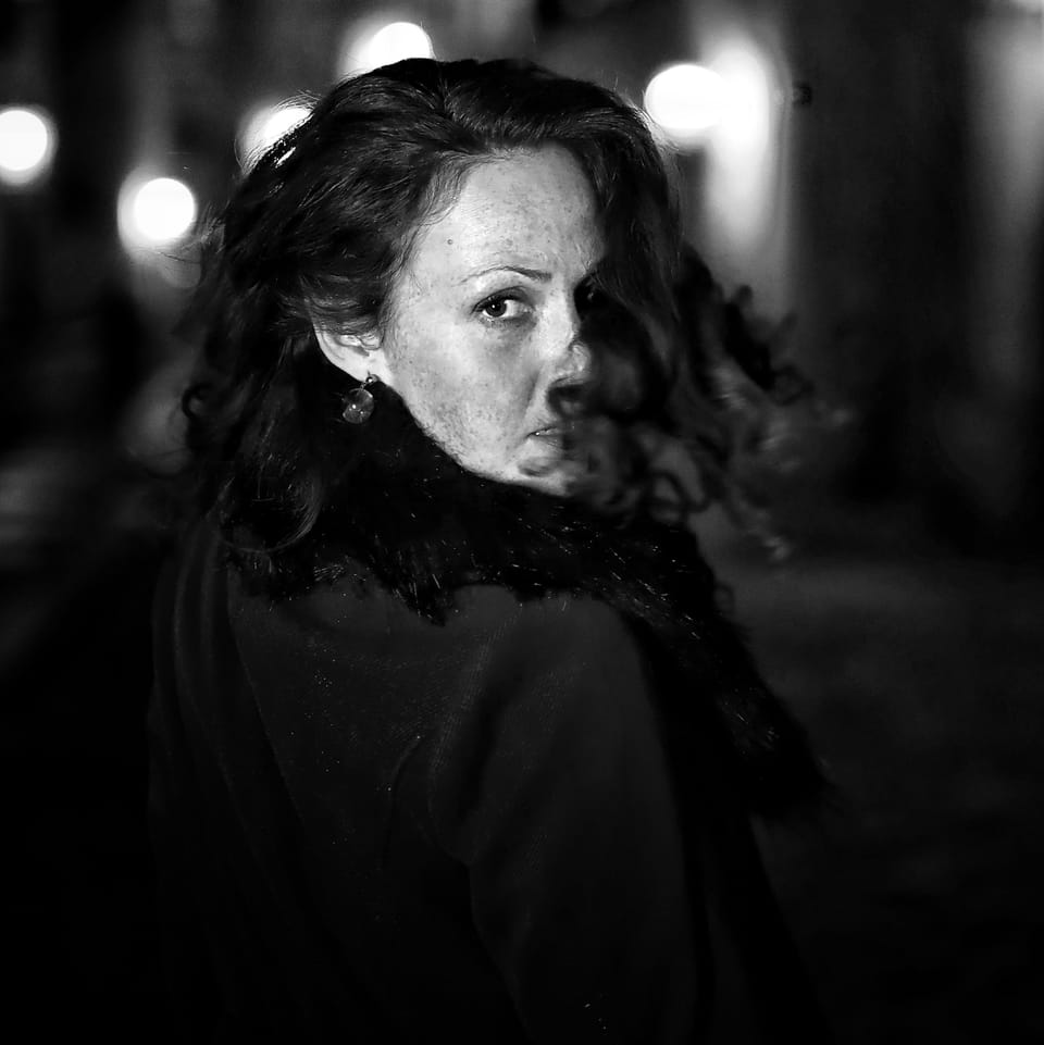 A woman in a dark coat glancing back over her shoulder, a night street behind