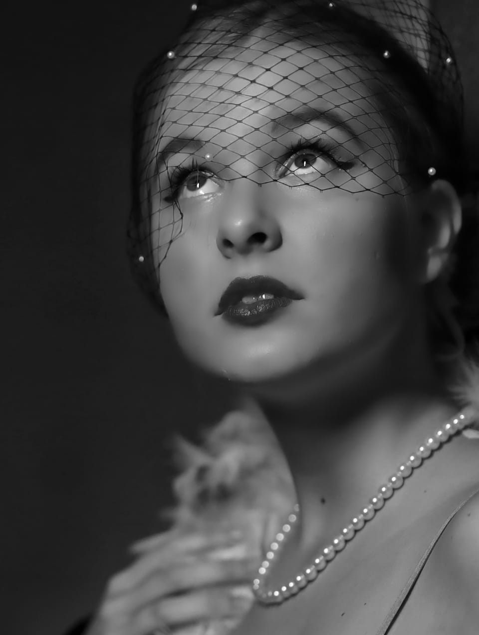 A woman in a net veil and pearls, her gaze lifted out of frame