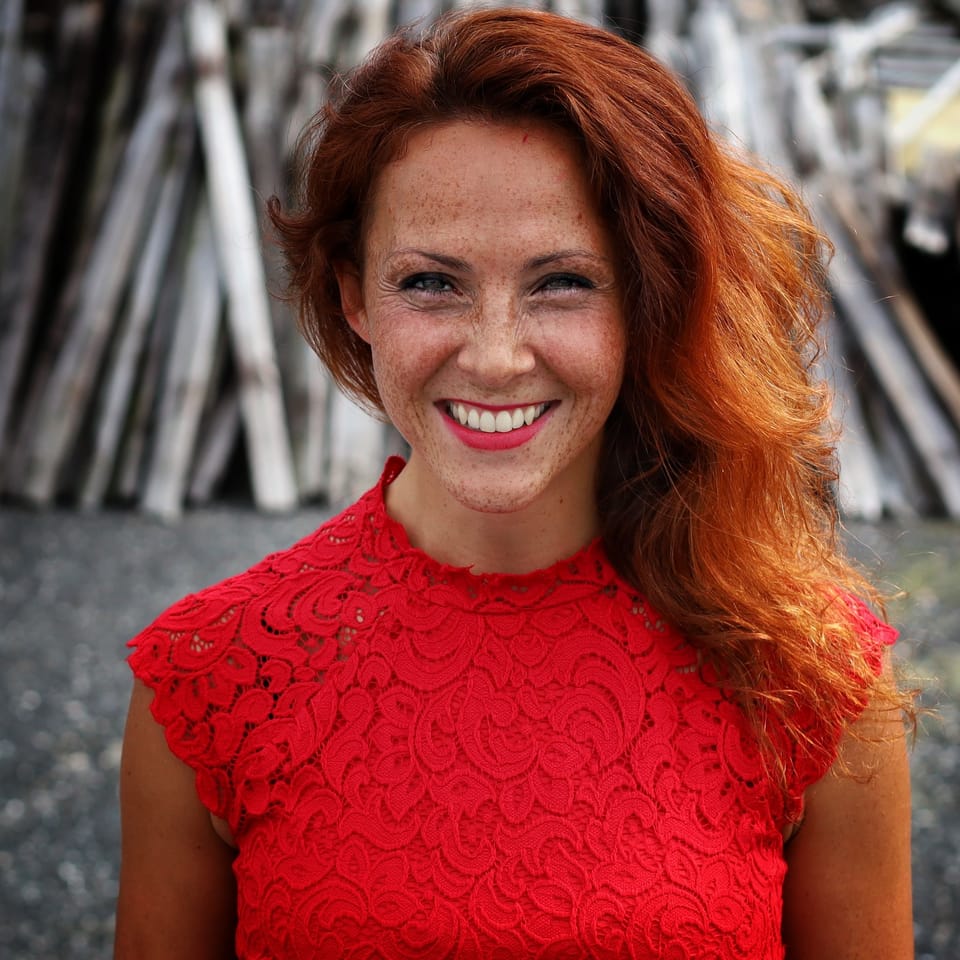 A smiling red-haired woman in a red lace top before a stack of timber
