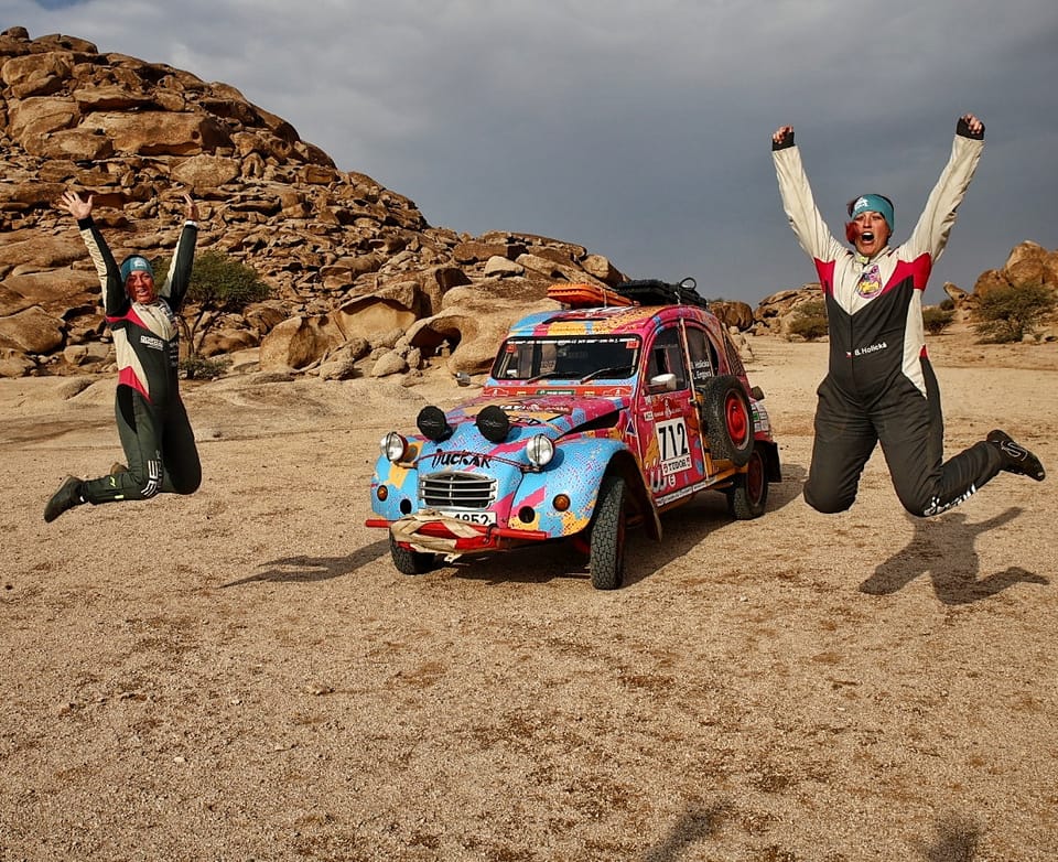 Two competitors leaping in the air beside their rally car in rocky terrain