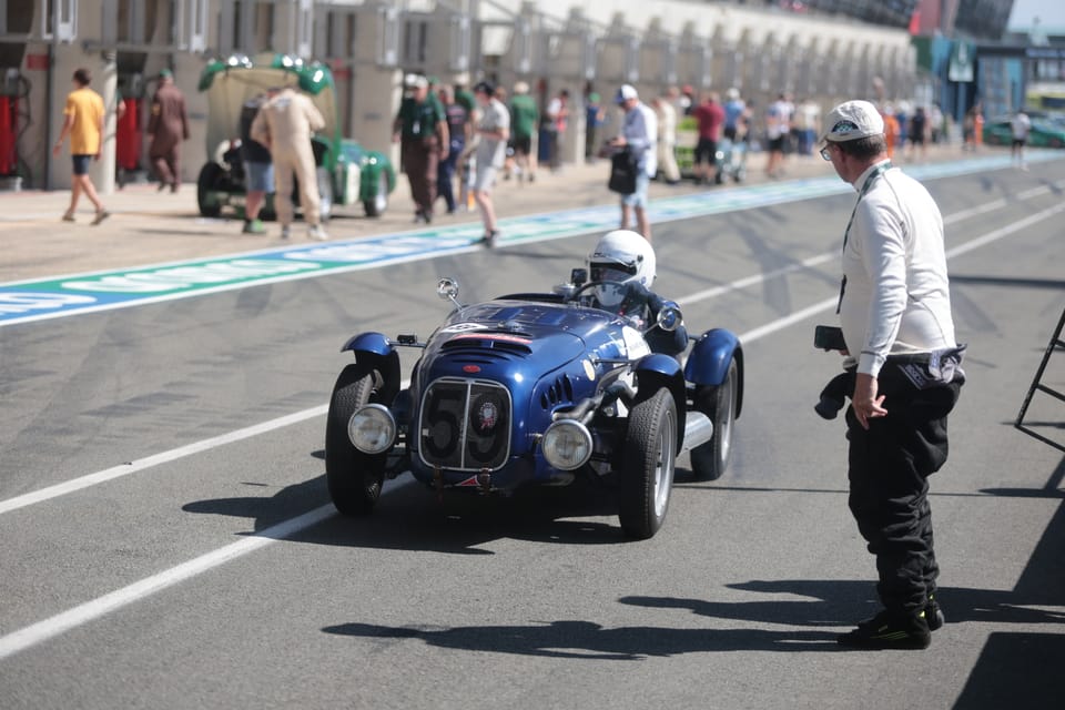 A blue historic roadster in the pits, a marshal standing alongside