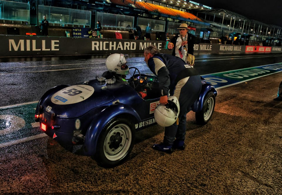 Wet pit lane: crew around a dark blue historic roadster, hoardings above