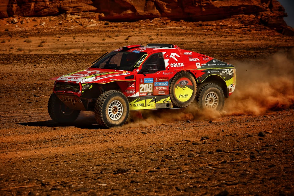 A buggy on stony flats below red rock, a plume of dust trailing behind