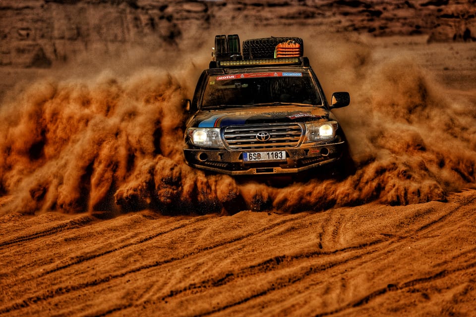 A Dakar rally car cutting through desert sand, a wall of dust rising behind it