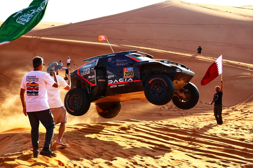 A Dakar car flying off a dune crest, photographers and marshals at the edge of the track