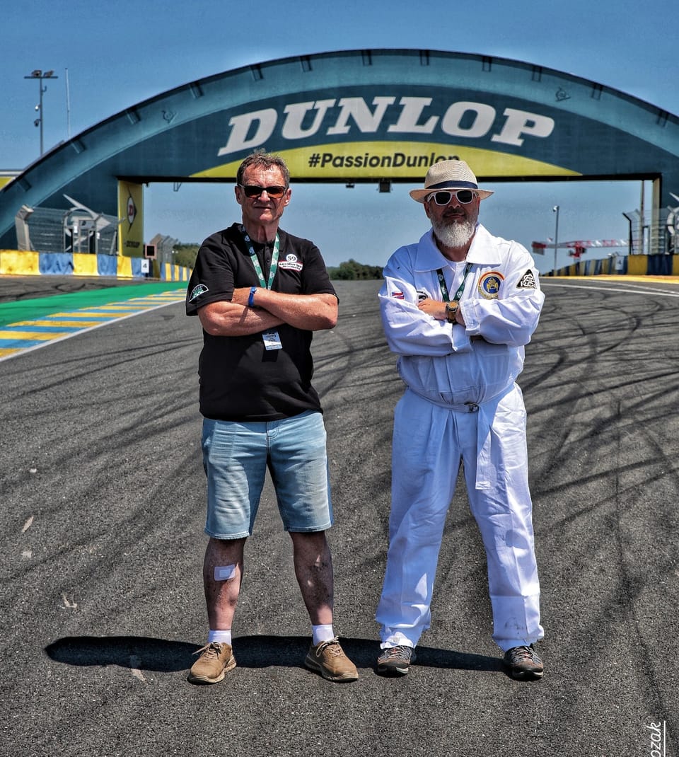 Two men beneath the arched bridge of the circuit, one in white racing overalls