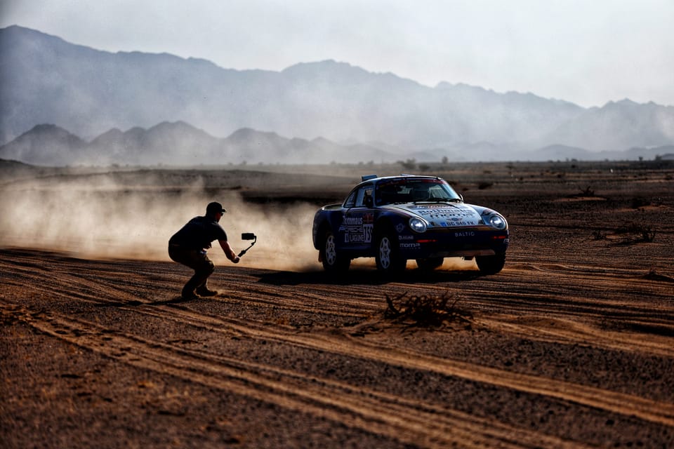 A photographer crouched in the sand shooting a passing car, a dusty plain stretching behind