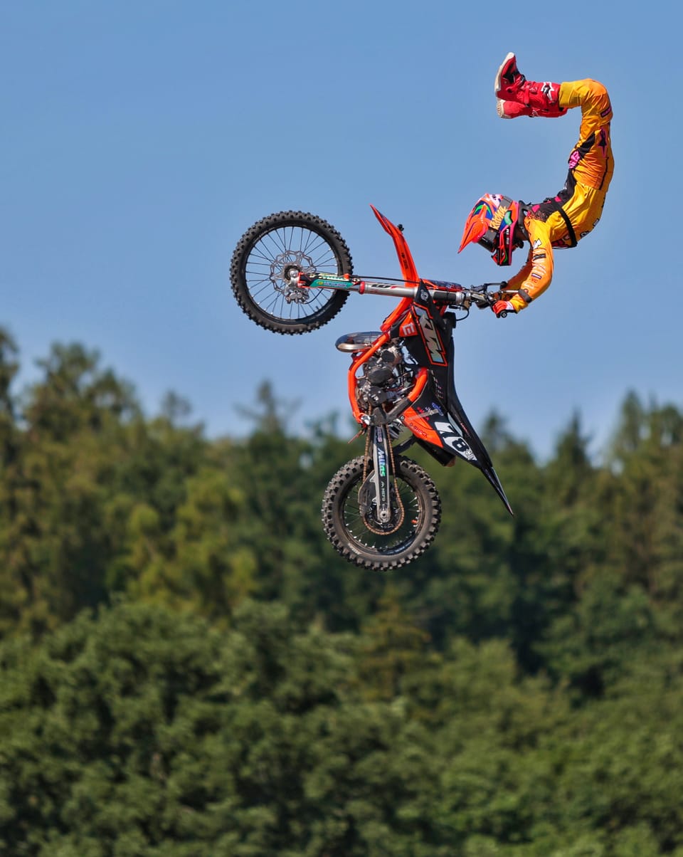 A freestyle motocross rider mid-jump, hands off the bars, stretched out above the treeline