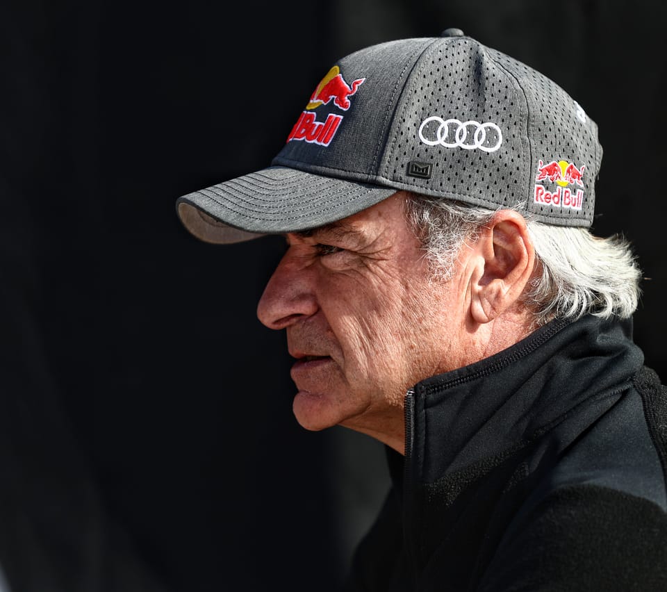A driver in profile against black, grey hair under a team cap