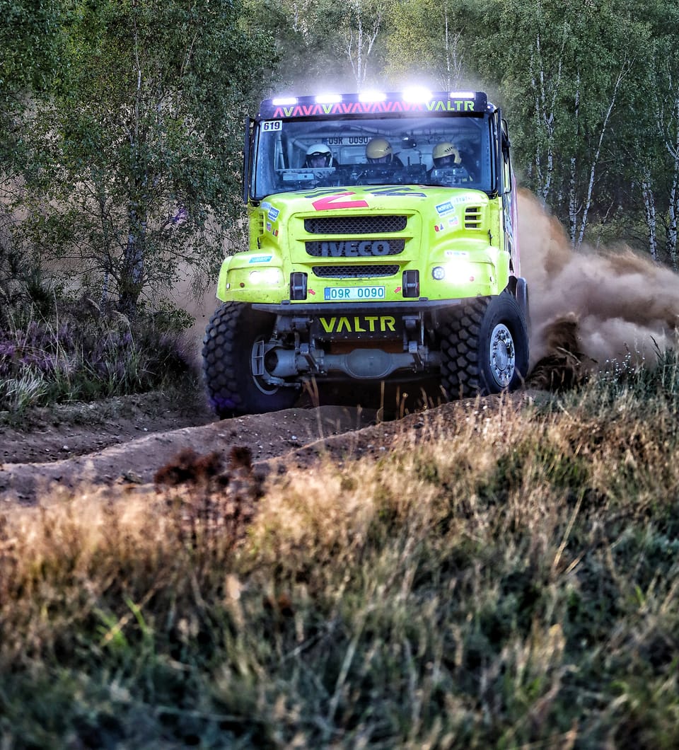 A yellow racing truck with its light bar lit on a forest track, birch and dry grass around