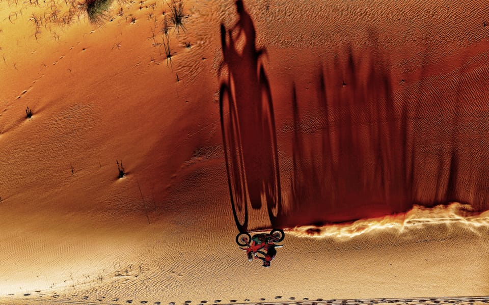 A motorcycle descending a vast orange dune, dragging a long track through the sand
