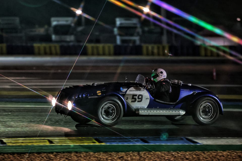 An open-top race car on a night circuit, floodlights breaking into coloured rays