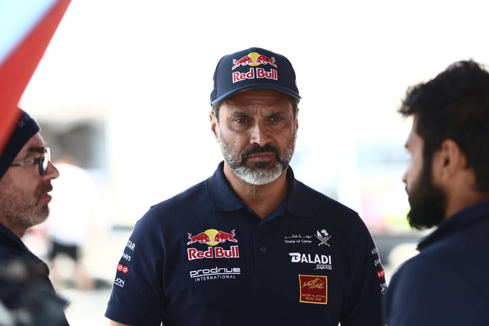 A paddock conversation: a man in team kit talking with two others, the bustle blurred around them