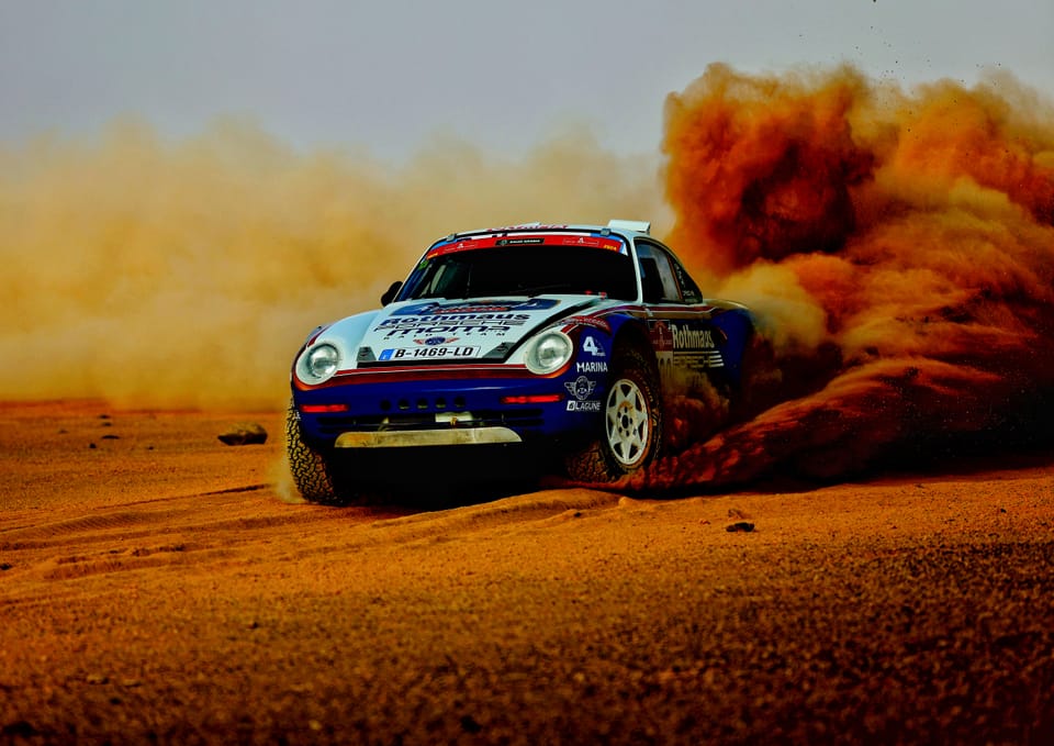 A historic Porsche in the desert, orange dust erupting behind its tail