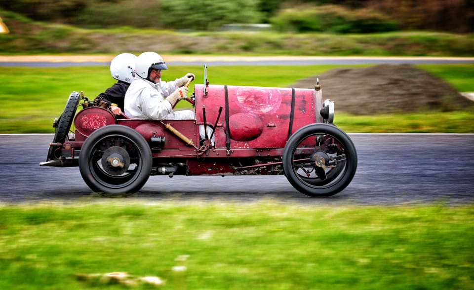 A pair in white helmets in an old open car, the background smeared by speed