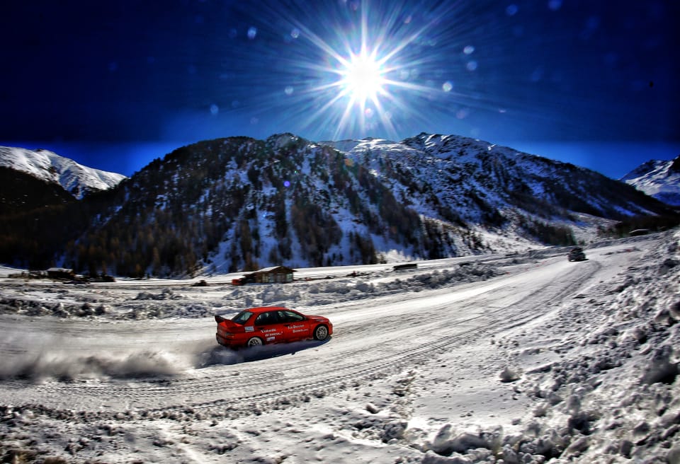 A rally car on a snowbound mountain road, the sun bursting into a hard star above the ridge