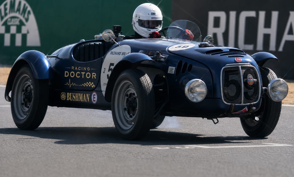 A dark blue historic roadster on the circuit, the driver in a white helmet above the low screen