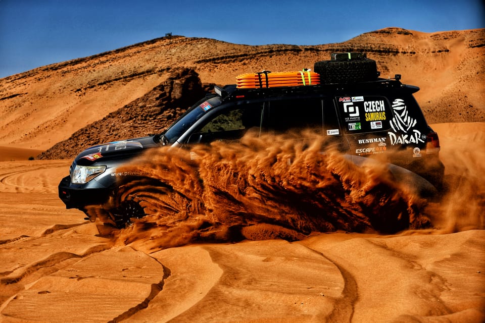 A rally raid car charging an orange dune, throwing out a fan of sand