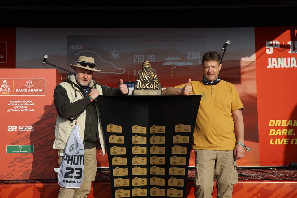 Two men giving a thumbs-up beside a displayed trophy, one with a camera and a press bib