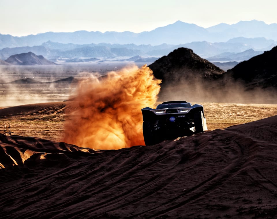 A car vanishing into an orange burst of dust, blue mountain outlines behind