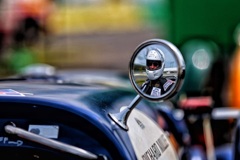 A helmeted driver mirrored in the round wing mirror of a historic car